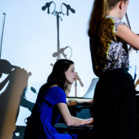 Two women, one in a blue dress playing piano, and one in a dark outfit playing violin
