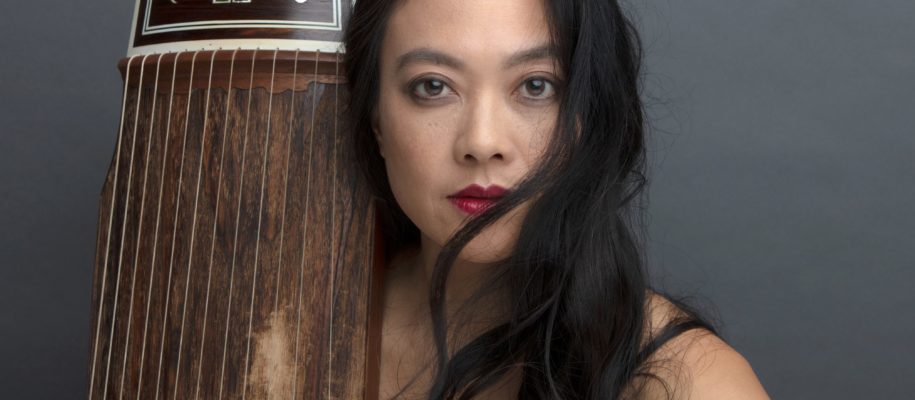 Jen Shyu with her hair in front of part of her face and a guzheng (classical Chinese zitrher) leaning against her right shoulder.