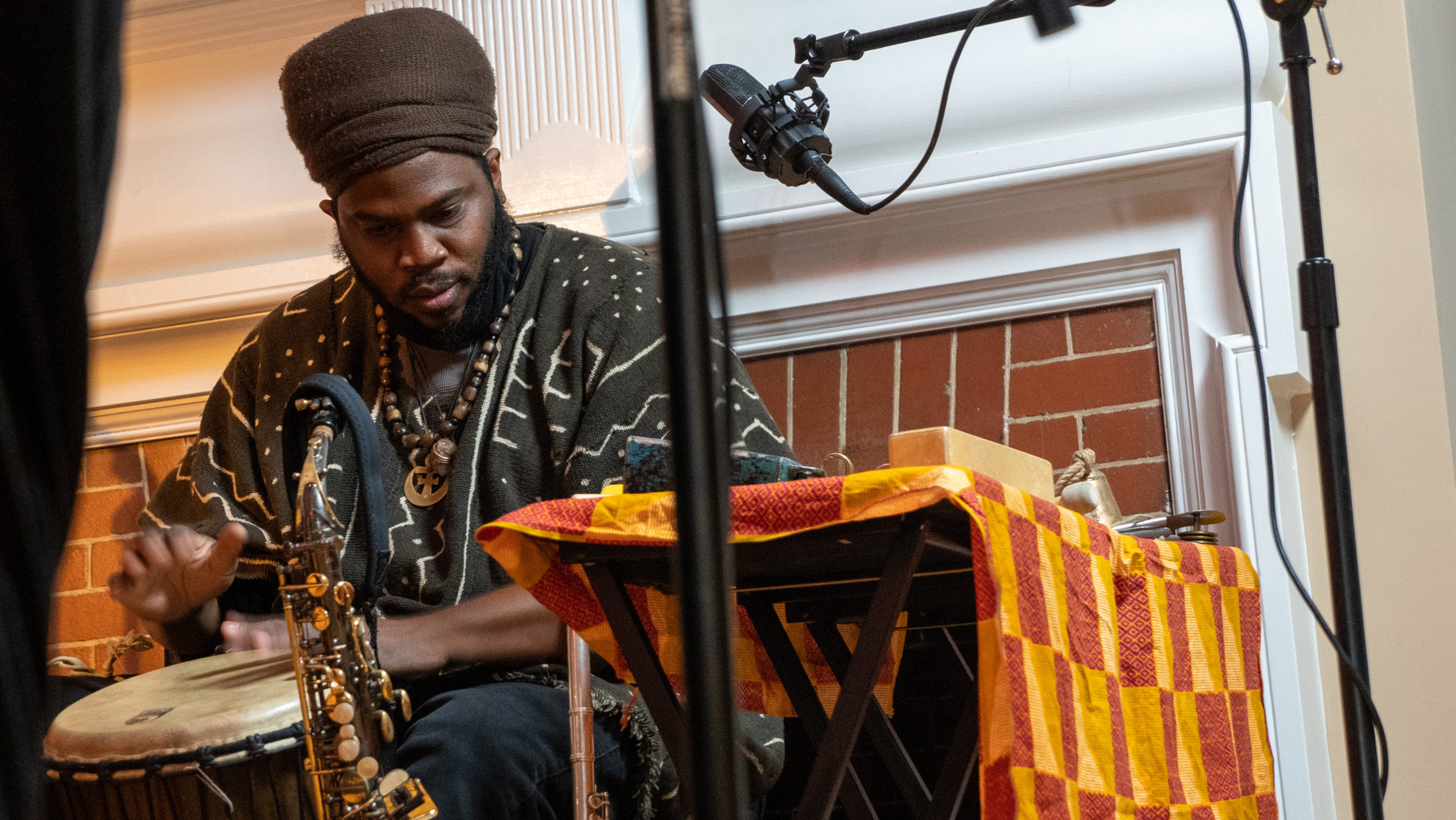 Jamal Moore in traditional African attire playing a hard drum as his saxophone sits on a stand nearby.