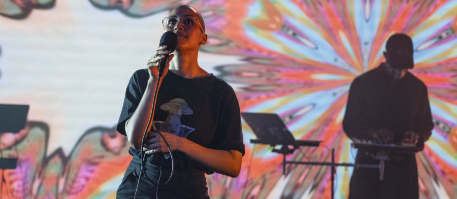 A colorful, flowery photo of a BIPOC woman waiting to sing and a man playing percussion/keyboard