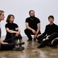 5 caucasian people sitting cross legged on the floor, looking at the person on the far right