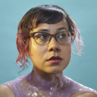 A woman with glasses and painted decoration around her neck against a teal background