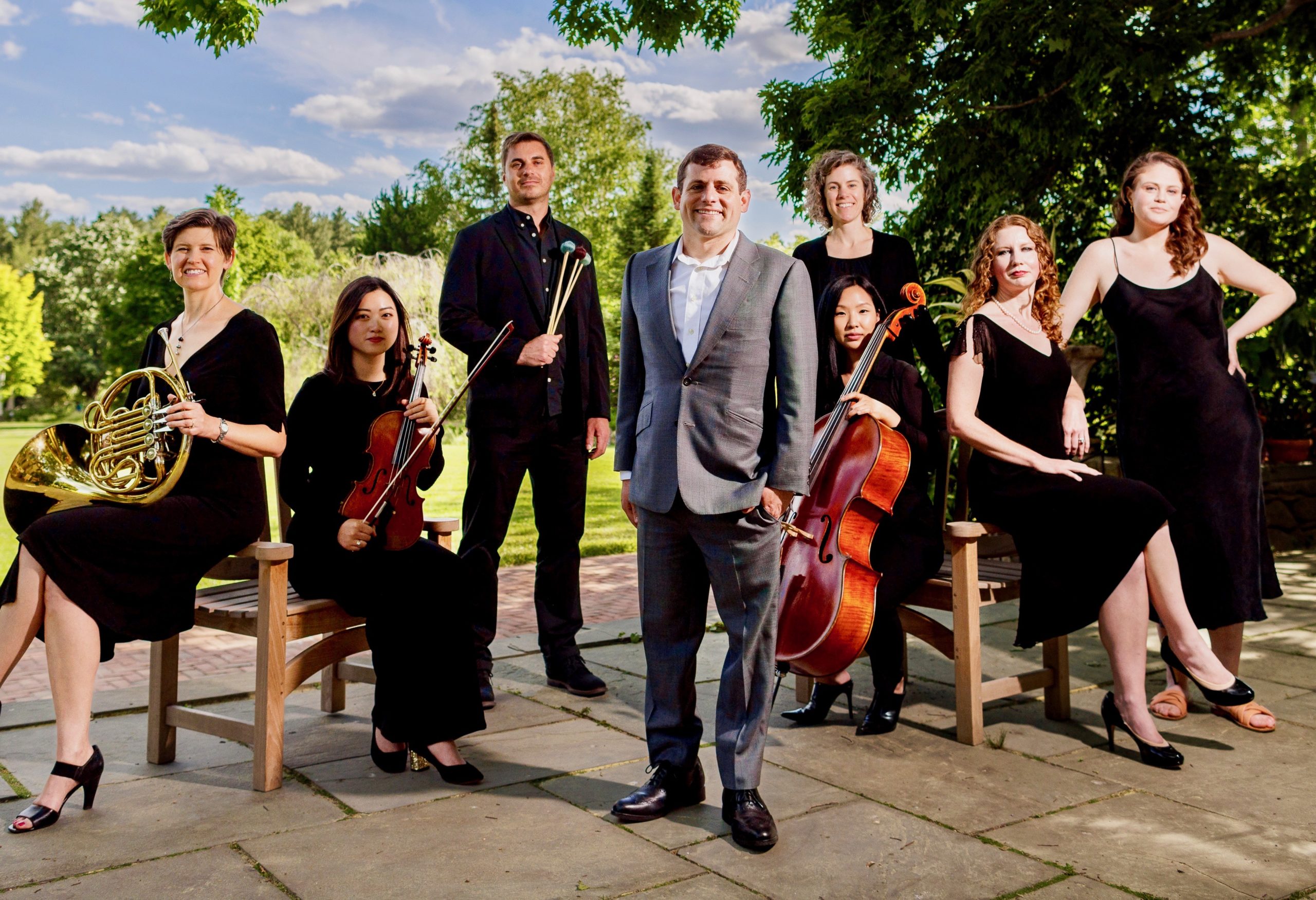 An outside photo of a group of musicians with various instruments with composer Oliver Caplan standing in the middle.
