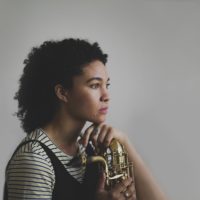 A woman posing with a baritone saxophone in a black-and-white striped shirt, head tilted
