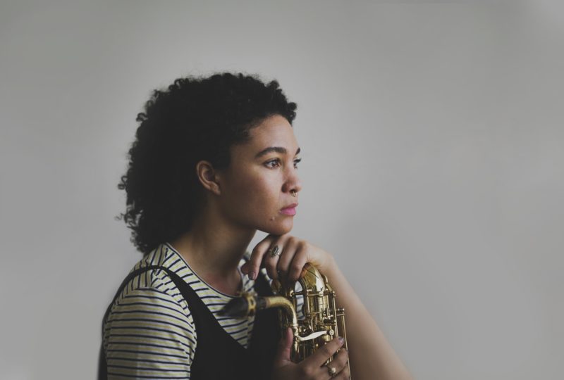 A woman posing with a baritone saxophone in a black-and-white striped shirt, head tilted