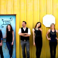 5 people posing in front of a yellow wall in dark clothing