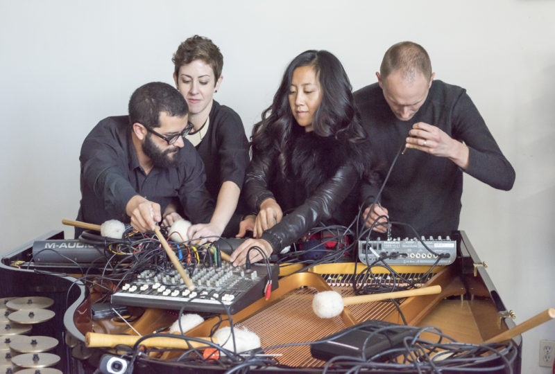 Four musicians crowd around electronics on a table, messing with knobs and wires