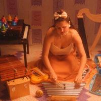 Alt Text / Image Description for alexa dexa and their toychestra: alexa dexa, in a silk gown with a white top and flowing peach skirtlength, kneels on the floor playing their toy chimes, Surrounding them from left to right are their 37-key Schoenhut toy piano, 20 chromatic desk bells, kalimba, alto xylophone, toy glockenspiel, tap-a-tune xylo-keyboard, and toy harp.